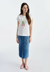 Biała koszulka z krótkim rękawem z haftem kwiatowym i różową wstążką, zestawiona z dżinsową spódnicą midi i jasnoróżowymi butami wsuwanymi.