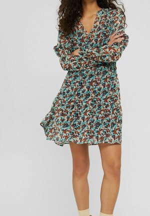 Kvinde med krøllet hår iført en langærmet, knælængde blomsterkjole i blå, brune og sorte toner, stående med armene krydsede.