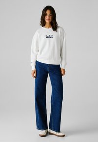 Sudadera blanca con el logo de "Red Bull Racing" en azul marino, combinada con jeans de pierna ancha en azul y zapatos color crema con suela marrón.