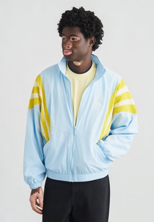 Jasnoniebieska kurtka na zamek z żółtymi paskami na rękawach, elastycznym pasem i małym logo na piersi. Noszona na żółtej koszulce.