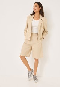 Blazer in lino beige abbinato a pantaloni corti plissettati coordinati, indossato sopra una maglietta bianca. Sneakers grigie completano l'outfit, aggiungendo un tocco casual.