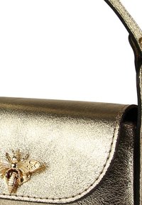 Goldfarbene metallic Handtasche mit glatter Textur, ausgestattet mit einem dekorativen Bienenemblem auf der Vorderklappe und einem passenden Tragegurt.