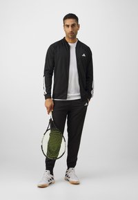Black athletic jacket with white stripes, matching black pants, and grey tennis shoes. Holding a tennis racket with a green and white string pattern.
