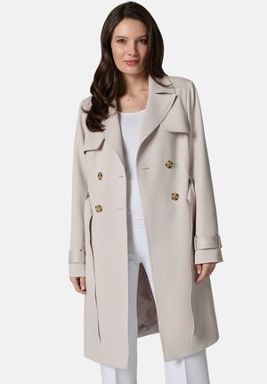 Kobieta ubrana w beżowy dwurzędowy trencz z guzikami w kolorze muszli żółwia, na białej bluzce i białych spodniach, stojąca na jednolitym tle.