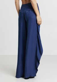 Persona che indossa pantaloni a vita alta e a gamba larga di colore blu navy con tasche welt posteriori, in piedi contro uno sfondo chiaro e neutro.