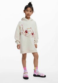 Robe sweat à capuche oversize couleur crème avec un graphique coloré du personnage Stitch sur le devant, associée à des sandales roses et noires et des chaussettes arc-en-ciel.
