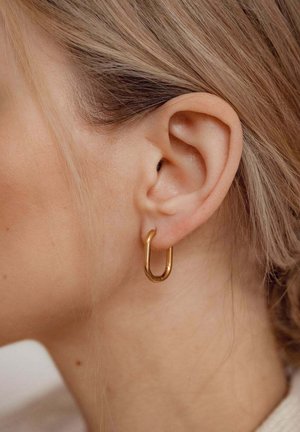 Gros plan sur l'oreille d'une personne à la peau claire et aux cheveux blonds, portant une petite boucle d'oreille épaisse en or.