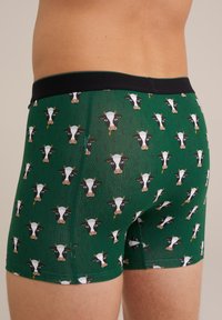 Zelene boksarice iz raztegljive tkanine z ponavljajočim se vzorcem risanih obrazov krav in črnim pasom.