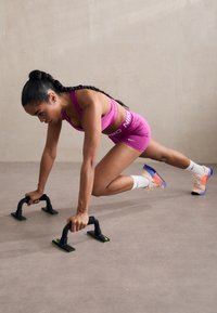 Barras de flexiones negras con acentos verdes sostienen a una persona en un sujetador deportivo rosa y pantalones cortos realizando un ejercicio sobre un suelo beige suave.