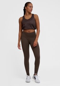 Brun cropped tanktop med geometriskt mönster, kombinerad med högmidjade bruna leggings. Modellen bär vita sportskor. Slät, åtsittande tyg.
