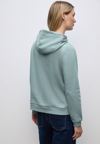 Felpa con cappuccio di colore azzurro chiaro, caratterizzata da una texture morbida, polsini e orlo a coste, progettata per una vestibilità rilassata. Vista posteriore su uno sfondo neutro.