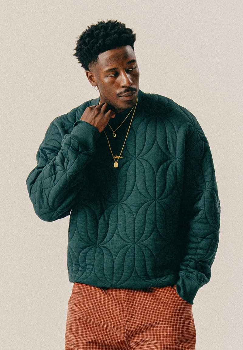 Domború sötétzöld pulóver, texturált mintával, bordázott mandzsettákkal és laza szabással, narancssárga kockás nadrággal párosítva.