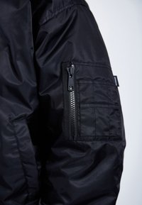 Black nylon jacket sleeve featuring a zippered pocket and square-shaped patch. The fabric appears smooth with a slight sheen.
