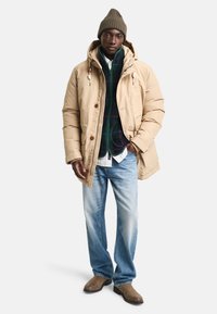 Beżowy długi parka z kapturem, zamkiem błyskawicznym i guzikami, nałożony na zielony sweter w wzory oraz jasnoniebieskie dżinsy. Brązowe buty dopełniają wygląd.