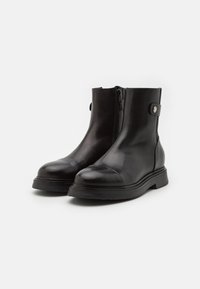 Bottines en cuir noir avec un bout arrondi, fermeture éclair sur le côté et une sangle décorative avec un bouton-pression en métal. Texture lisse et semelle robuste.