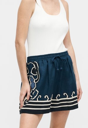 Γυναίκα που φοράει λευκό ριμπ ζιπ-παντόφλα και σκούρα μπλε σορτς με κρεμ χρώματος λουλουδάτη κέντημα και κορδόνι στη μέση.