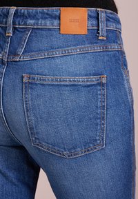 Vaqueros de mezclilla azul con una textura descolorida, que presentan un detalle de costura naranja y un parche de cuero marrón claro etiquetado como "CLOSED" en la cinturilla.