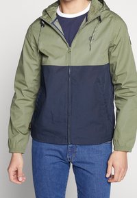 Hoodiejacka med grön övre del och mörkblå nedre del, tillverkad av lätt material. Har elastiska muddar och sidofickor.