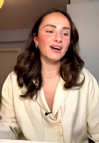 Jonge vrouw met gekruld donker haar en lichte blouse spreekt binnenshuis met een microfoon vastgeklemd aan haar overhemdkraag.