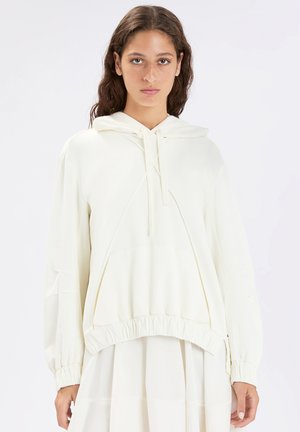 Donna con capelli ricci che indossa una felpa bianca oversize con cappuccio, tasca a marsupio ed orlo elastico, su uno sfondo chiaro e uniforme.