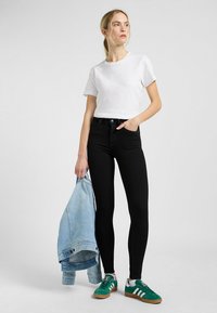 Črne visokotlačne kavbojke s tesnim krojem, v kombinaciji z belo kratkonožno majico in zelenimi športnimi čevlji. Lahka denim jakna ji je prevrnjena čez roko.