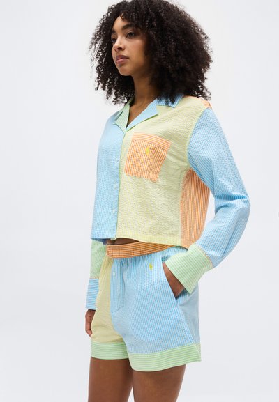 Γυναίκα που φοράει πολύχρωμο πουκάμισο με σχέδιο κομμάτια ραμμένα μαζί και ταιριαστό σορτς με τσέπες, στέκεται μπροστά σε ένα απλό φόντο.