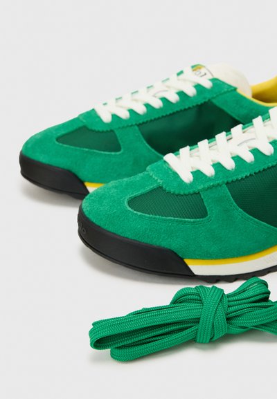 Zelené semišové tenisky s sieťovinou a bielymi šnúrkami, s čiernou gumovou podrážkou a žltými akcentmi. Obsahuje aj ďalšiu zelenú šnúrku.