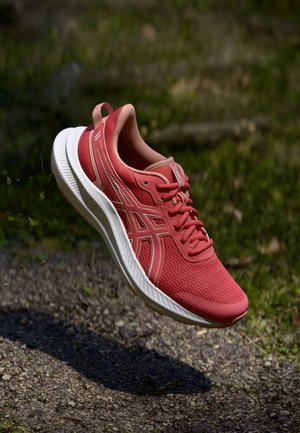 Red running shoe with white sole suspended above gravel path, surrounded by blurred green grass background.