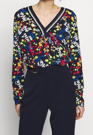 Frau trägt eine marineblaue Bluse mit langen Ärmeln und V-Ausschnitt, verziert mit einem bunten Blumenmuster, sowie eine hochtaillierte marineblaue Hose.