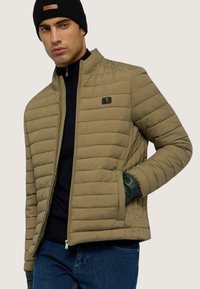 Zateplená olivově zelená bunda s vysokým límcem, zapínáním na zip a malým logem na hrudi. Nositá přes námořnický svetr a modré džíny.