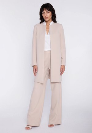 Kobieta stojąca w beżowym długim żakiecie i szerokich spodniach, białej bluzce oraz białych sandałach z odkrytymi palcami na jednolitym tle.