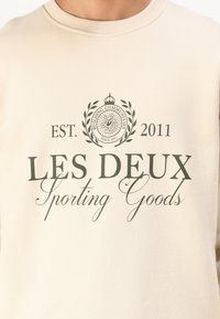 Beige sweater met een circulair logo met een kroon en laurierontwerp, gedrukt in donkergroen. Tekst bevat "LES DEUX" en oprichtingsjaar "2011."