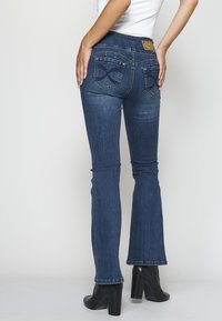 Blå utsvängda jeans med medelhög midja, två bakfickor med dekorativ sömnad och subtila blekningsdetaljer. Svarta klackskor visas.