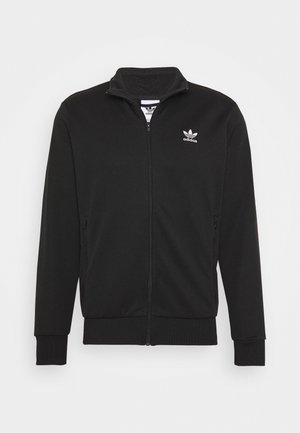 Man draagt zwarte trainingsbroek met witte strepen, zwart jack met rits, wit T-shirt en zwarte hoge sneakers, staat met handen in de zakken.