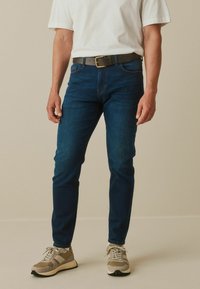 Jeans azul oscuro de corte ajustado, combinados con una camiseta blanca y zapatillas beige. Tela texturizada notable y un cinturón marrón con hebilla dorada.