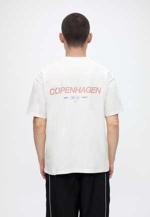 Άτομο που φοράει λευκό μπλουζάκι με κόκκινο κείμενο "COPENHAGEN" και μικρότερο μπλε κείμενο στην πλάτη, συνδυασμένο με μαύρο παντελόνι με λευκές ρίγες.