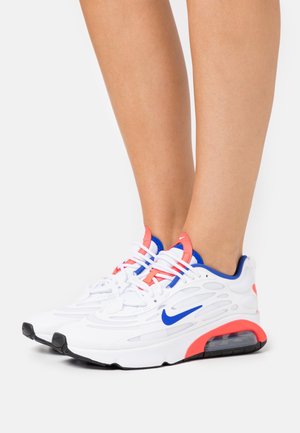 Sneakers atletiche bianche con logo Nike blu, accenti corallo e cuscinetto ad aria visibile, indossate a piedi nudi su uno sfondo semplice.