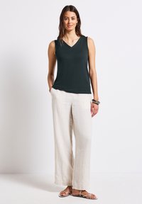 Halenka bez rukávů ve tmavě zelené barvě s výstřihem do V, spárovaná se světlými béžovými širokými lněnými kalhotami. Modelka stojí s jednou rukou v kapse a má na sobě ploché sandály.