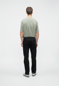 Camiseta corta de manga verde claro, jeans negros y zapatos negros con detalles grises; visto desde la parte de atrás contra un fondo liso.