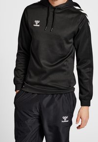 Sudadera negra con una textura suave, que presenta un logotipo blanco en el pecho y rayas contrastantes en las mangas, combinada con pantalones negros.