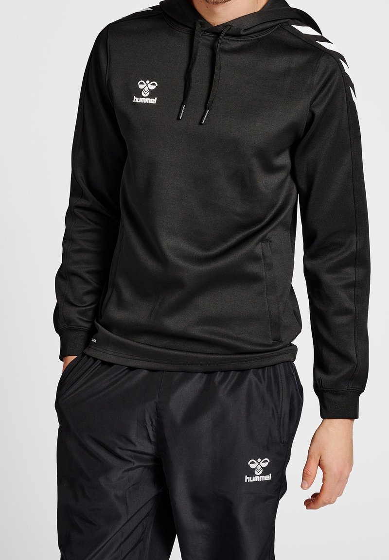 Sudadera negra con una textura suave, que presenta un logotipo blanco en el pecho y rayas contrastantes en las mangas, combinada con pantalones negros.