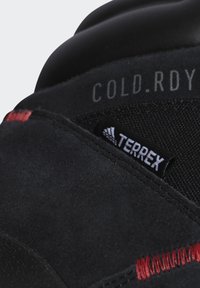 Czarny but sportowy z zamszową cholewką, z czerwonymi przeszyciami, teksturowanym panelem materiałowym oraz etykietą "COLD.RDY". Zawiera oznaczenie "TERREX".
