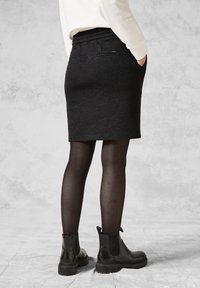 Černá sukně ke kolenům s elastickým pasem, dvěma zadními kapsami a hladkým povrchem, kombinovaná s černými kotníkovými botami a neprůhlednými punčochami.