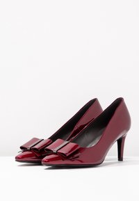 Escarpins en cuir verni bordeaux avec des pointes et un détail en nœud à l'avant. Les talons aiguilles offrent une silhouette élégante. Texture lisse.