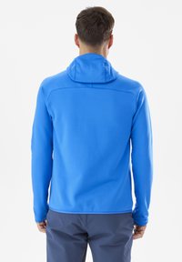 Homme portant une veste à capuche bleu vif et un pantalon noir, standant de dos contre un fond blanc uni.