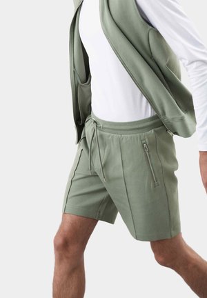 Mann trägt salbegrüne Zip-up-Weste und passende Shorts mit Reißverschlusstaschen, kombiniert mit einem weißen Langarmshirt, beschnittener Ausschnitt.