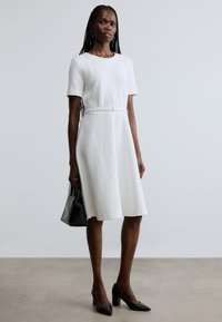 Robe blanche à manches courtes avec un motif texturé, une taille cintrée et une jupe évasée. Associée à des chaussures à talons hauts noires et un sac à main noir.