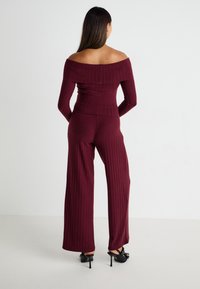 Burgundy csíkos, váll nélküli felső, amely széles szárú nadrággal van párosítva, textúrált anyaggal és formázott kialakítással. Fekete magassarkú cipő.