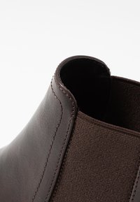 Botín de tobillo de cuero marrón con paneles laterales elásticos. La parte superior presenta una textura suave con un detalle de costura en contraste.