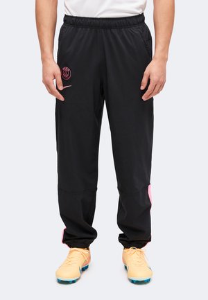 Personne portant un pantalon de sport noir avec un logo rose du Paris Saint-Germain et des chaussures de football pêche avec des crampons bleus, debout devant un fond uni.